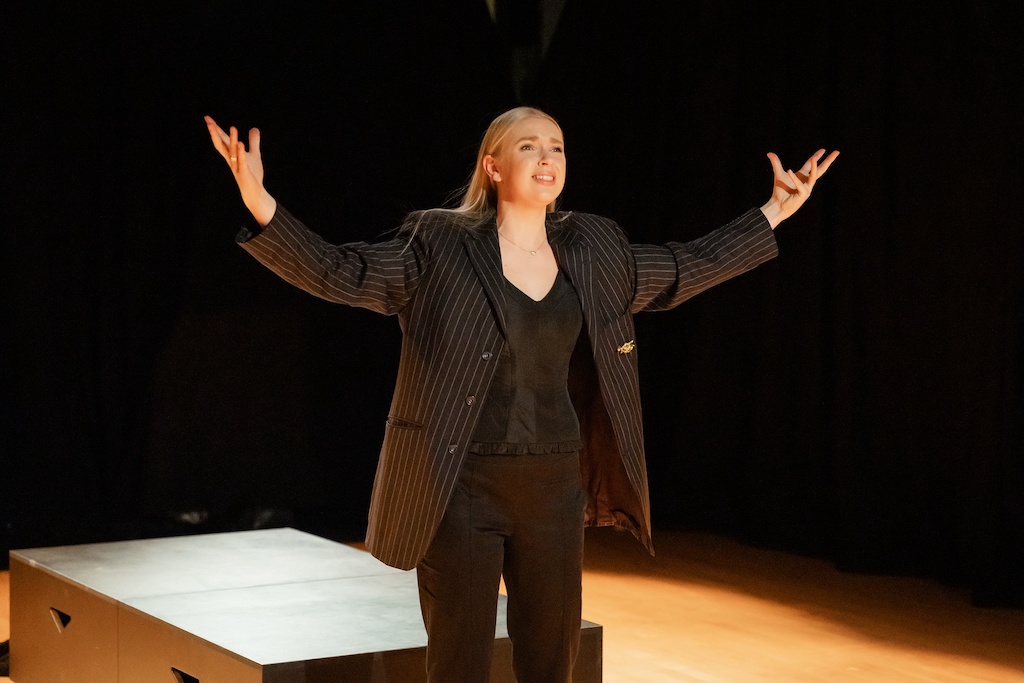 An actor wearing an all-black outfit stands on stage in mid-performance with arms outstretched.